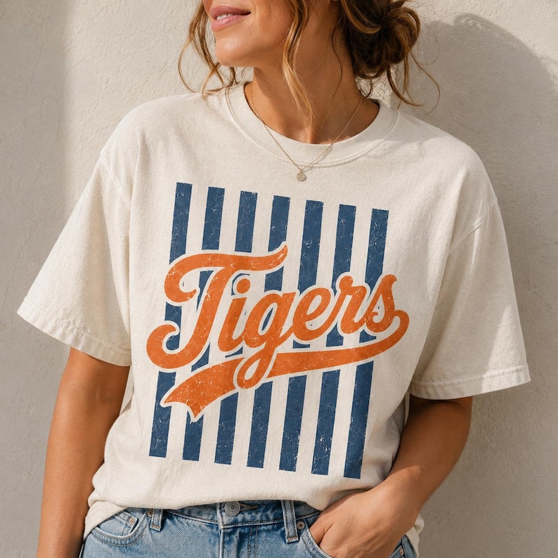 May include: A cream-colored short-sleeve t-shirt featuring a graphic design with vertical navy blue stripes and the word Tigers written in a large, distressed orange script font across the center. The shirt has a crew neckline and a relaxed fit.