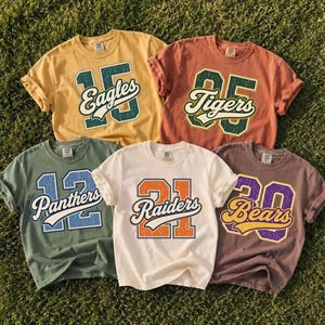 May include: Five short-sleeve crew neck t-shirts in yellow, terracotta, sage green, cream, and mauve. Each shirt features a large varsity-style number graphic with a team name written in script across the center: Eagles 15, Tigers 05, Panthers 12, Raiders 21, and Bears 20.