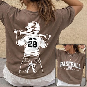 May include: Brown t-shirt with a white graphic of a baseball player holding a bat, the name "Thomas" and the number "28" on the back. A smaller image shows a brown t-shirt with the words "Baseball mom" and a baseball.