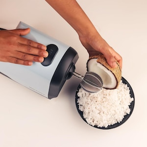 May include: An electric coconut grater in use. The light blue and black appliance is grating a coconut half over a black bowl filled with shredded coconut. The grater has a stainless steel blade.