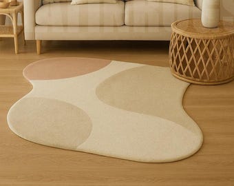 Könnte beinhalten: Ein abstrakt geformter Teppich in neutralen Tönen von Beige, Hellbeige und Rosé. Der Teppich hat eine weiche, organische Form mit abgerundeten Abschnitten. Er liegt auf einem hellen Holzboden, mit einem beigen Sofa und einem gewebten Beistelltisch im Hintergrund.