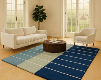 May include: A modern living room scene featuring a cream-colored sofa and armchair, a dark brown round coffee table, and a blue and gray striped rug. The rug has a geometric pattern with white stripes. The room has large windows and a potted plant.