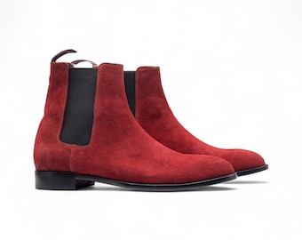 May include: A pair of red suede Chelsea boots. The boots have a classic design with elastic side panels and a pull tab at the back. The soles are black, and the boots are set against a white background.
