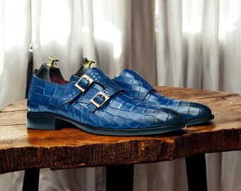 May include: A pair of blue leather double monk strap shoes with a textured, crocodile-like pattern. The shoes have gold-colored buckles and are displayed on a wooden surface. Shoe trees are inserted inside the shoes.