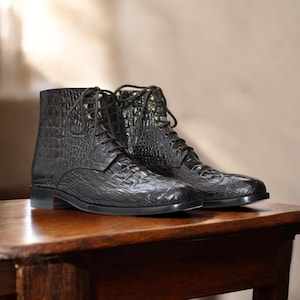 May include: A pair of dark brown leather boots with a crocodile skin texture. The boots have laces, metal eyelets, and a low heel. They are displayed on a wooden surface.