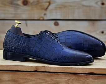 May include: A pair of blue leather dress shoes with a textured, crocodile-like pattern. The shoes have a classic lace-up design and a sleek, polished appearance. The sole of one shoe is visible, showcasing a grid pattern.