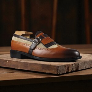 May include: A brown leather shoe with a decorative fringe and buckle closure. The shoe features a two-tone design with a lighter tan suede panel and a darker brown leather strap. The sole is black.