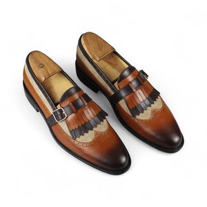 May include: A pair of brown leather loafers with decorative fringe and a buckle closure. The shoes have a two-tone design with tan and dark brown accents. Wooden shoe trees are inserted inside the shoes.