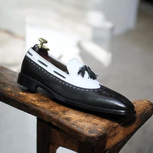 Side profile showing elegant black & white design with stylish tassel detail and sleek slip-on style.