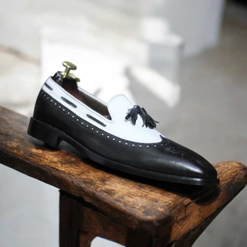 Side profile showing elegant black & white design with stylish tassel detail and sleek slip-on style.