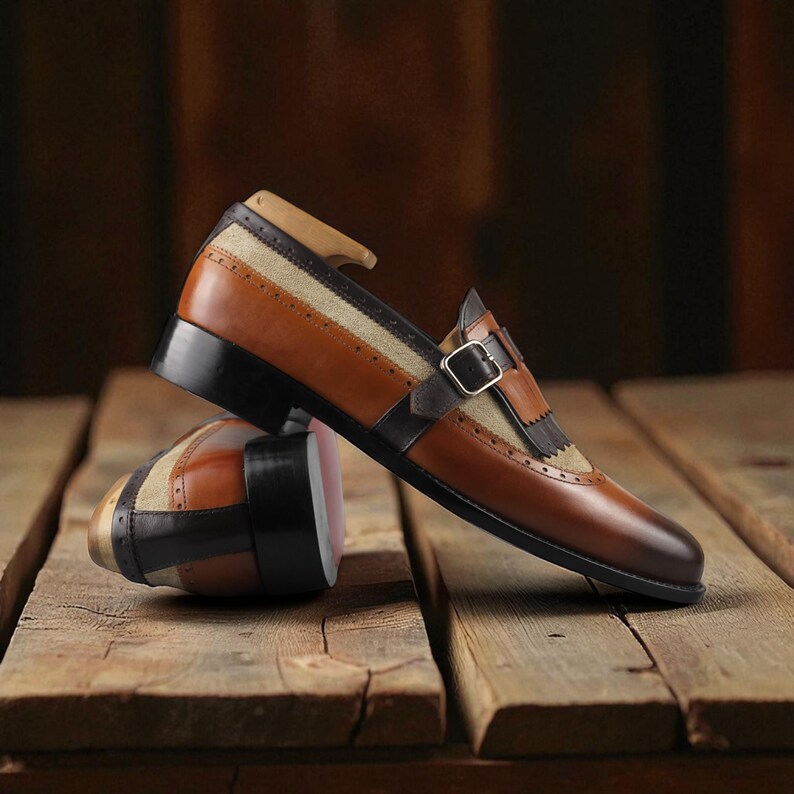 May include: A pair of brown and tan leather dress shoes with a buckle closure. The shoes have a unique design with a combination of smooth and textured leather, and are set on a wooden surface.