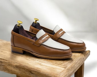 May include: A pair of two-tone leather loafers. The shoes are brown with white accents on the vamp and saddle. The shoes are displayed on a wooden surface, with shoe trees inserted. The loafers have a classic, polished appearance.