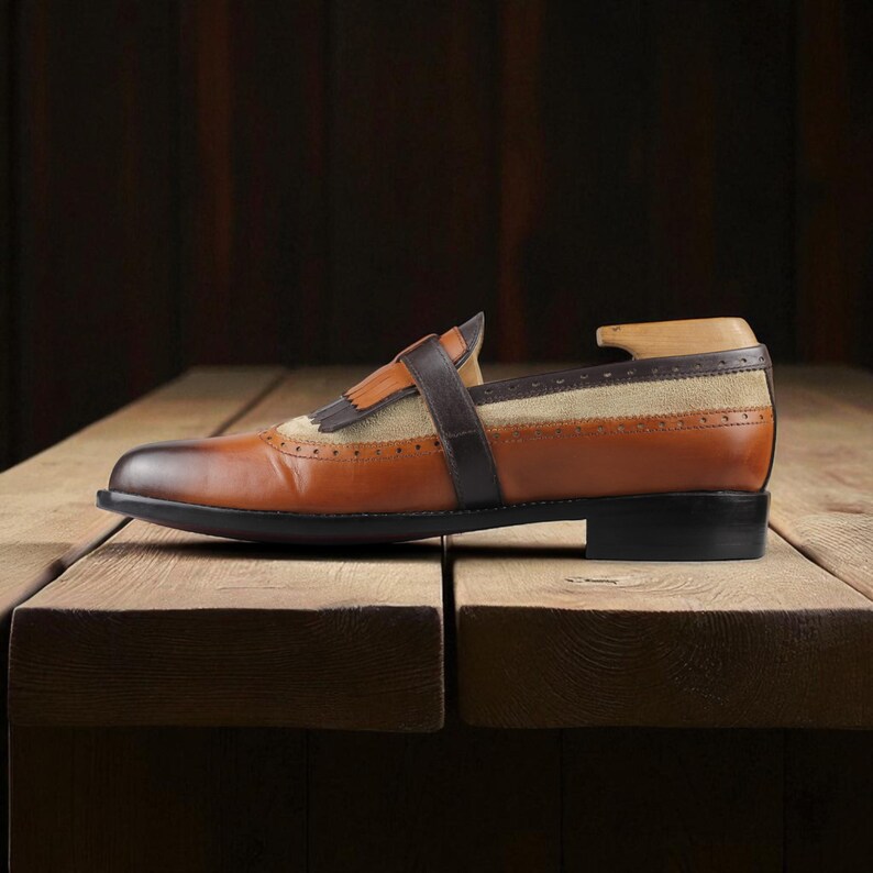 May include: A brown and tan leather loafer with a fringe detail and a strap across the top. The shoe has a black sole and is displayed on a wooden surface. The shoe has a wooden shoe tree inside.