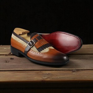 May include: A pair of brown leather dress shoes with a unique design. The shoes feature a tan fabric panel, a buckle strap, and decorative fringe. The soles are a deep red color. The shoes are displayed on a wooden surface.