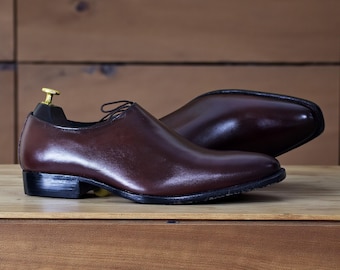 May include: A pair of brown leather dress shoes with a low heel and a sleek, polished finish. The shoes are displayed on a wooden surface, showcasing their elegant design and craftsmanship. The laces are thin and dark.