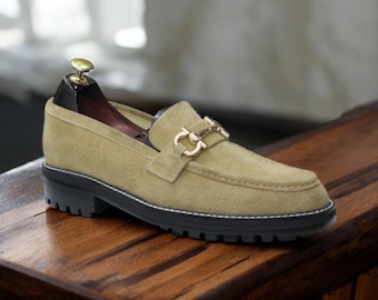 May include: A tan suede loafer with a gold-colored buckle and a black lug sole. The shoe has a classic design with a rounded toe and a stitched welt. The shoe is approximately 1.5 inches high.