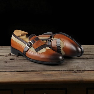 May include: A pair of brown leather dress shoes with a buckle closure and decorative fringe. The shoes have a two-tone design with tan and brown suede accents. The shoes are displayed on a wooden surface.