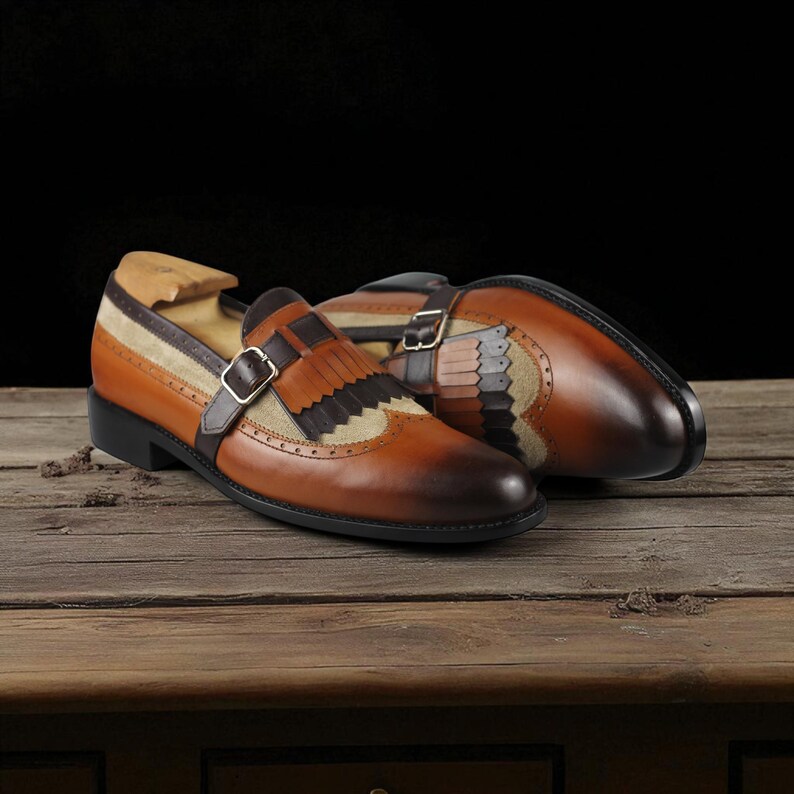 May include: A pair of brown leather dress shoes with a buckle closure and decorative fringe. The shoes have a two-tone design with tan and brown suede accents. The shoes are displayed on a wooden surface.