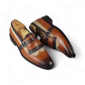 May include: A pair of brown leather loafers with decorative fringe and buckle details. The shoes feature a two-tone design with tan and dark brown accents. The soles are black, and the shoes are displayed on a white background.