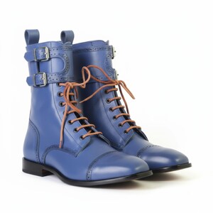 May include: A pair of blue leather boots with brown laces and silver buckles. The boots have a classic design with a cap toe and decorative perforations along the seams. The boots are set against a white background.