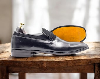 May include: A black leather loafer with a suede accent and white stitching. The shoe is displayed with a wooden shoe tree and a yellow insole. The shoe is on a wooden surface.
