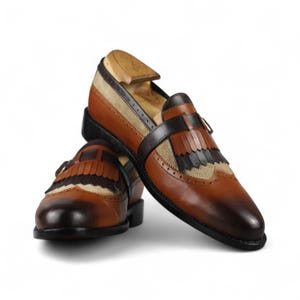 May include: A pair of brown leather loafers with decorative fringe and a contrasting tan suede panel. The shoes have a classic design with a dark brown strap and a wooden shoe tree inside.