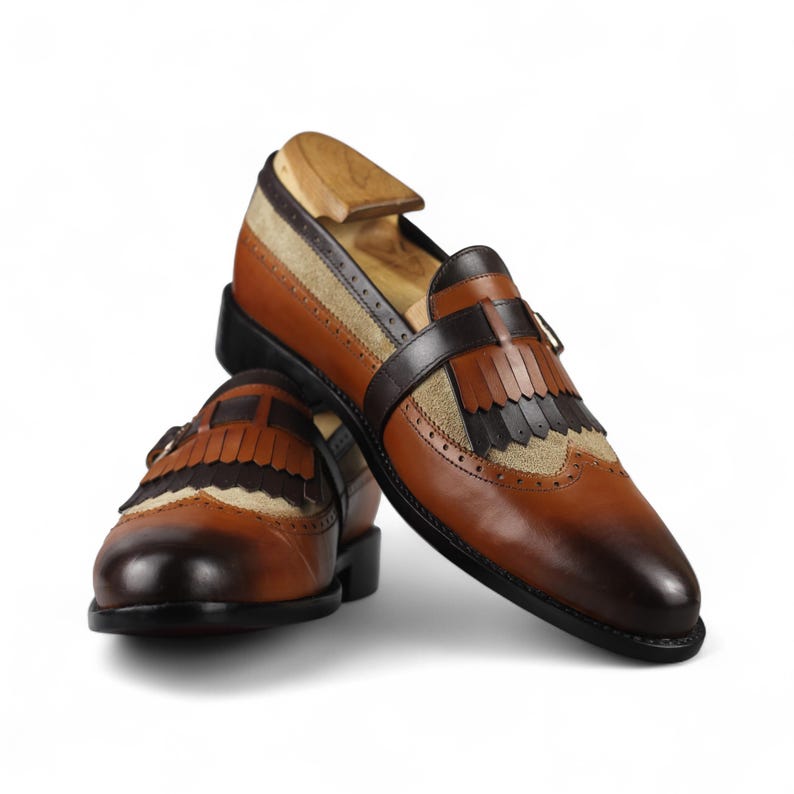 May include: A pair of brown leather loafers with decorative fringe and a contrasting tan suede panel. The shoes have a classic design with a dark brown strap and a wooden shoe tree inside.