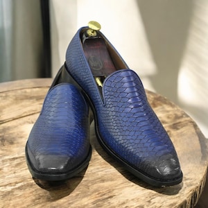 Pode incluir: Um par de mocassins de couro azul com textura de pele de cobra. Os sapatos têm um efeito gradiente, passando de um tom mais escuro na ponta para um azul mais claro. Os sapatos são exibidos em uma superfície de madeira.