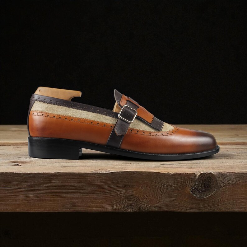 May include: A single brown leather shoe with a black sole, buckle, and tan fabric panel. The shoe is displayed on a wooden surface against a dark background. The shoe has a classic design with a modern twist.