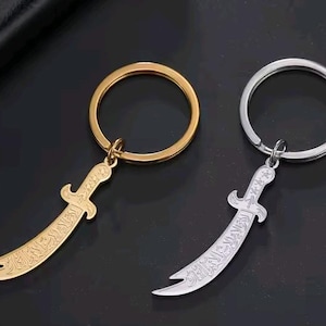 May include: Two keychains with a curved sword pendant. One keychain is gold-colored, the other is silver-colored. Both pendants have engraved details and are attached to a round key ring. The background is dark.