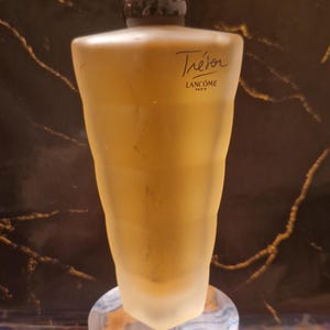 Puede incluir: Un frasco de perfume dorado con un tapón transparente facetado. La botella tiene la palabra "Trésor" en escritura. La botella se exhibe sobre un soporte cilíndrico decorativo con remolinos azules y blancos.