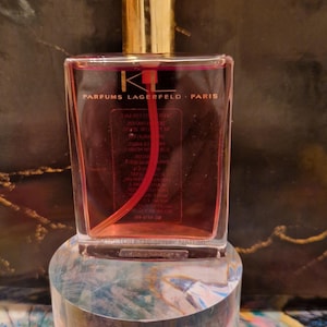 以下が含まれることがあります： 金色のキャップと透明でピンク色の液体が入った長方形の香水瓶。ボトルには「PARFUMS LAGERFELD PARIS」の文字が印刷されています。ボトルは透明な円筒形のスタンドに展示されています。