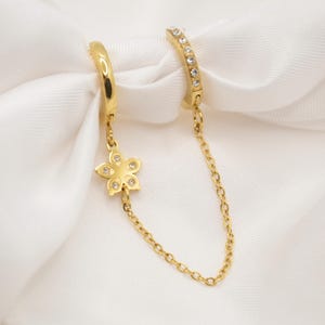 Peut inclure: Boucles d'oreilles doubles dorées. Une boucle d'oreille créole est reliée par une chaîne délicate à une seconde boucle d'oreille créole ornée de petits cristaux transparents. Un charm en forme de fleur avec des cristaux transparents est suspendu à la première boucle.