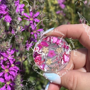 May include: A circular resin piece containing pink abalone shell fragments, crushed glass, and dried flowers including Pyracantha blossom, Japonica blossom, and lace flower. The piece features white line art of a dolphin, octopus, and shell.