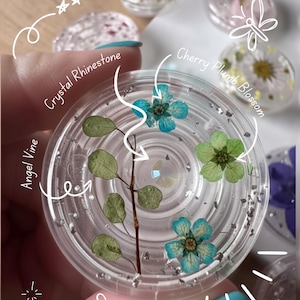 Könnte beinhalten: Ein klarer, runder Handyhalter mit eingebetteten getrockneten botanischen Elementen, darunter eine braune Engel-Rebe, zwei blaue Blüten und eine grüne Kirschblüte. Ein zentraler Kristall-Strassstein ist von Silberflocken umgeben.