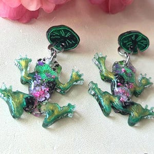 May include: A pair of whimsical frog earrings. Each earring features a green frog with gold accents and a lily pad stud. The frogs are filled with colorful floral details. The earrings are set against a soft, off-white background.