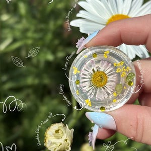Op de afbeelding: Een heldere ronde harsbedel met een geperste tuinmargriet, kleine gele kantbloemen en gele kristallen. De bedel is voorzien van tekstlabels, waaronder Shashta Daisy in our flower gardens, Lace Flower, Garden Daisy en Yellow Crystal.