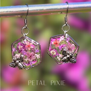 May include: A pair of hexagonal earrings featuring clear resin filled with small pink and purple pressed flowers. Each earring is accented with a silver-toned metal bee charm attached to the bottom corner. The earrings hang from silver-toned hooks.