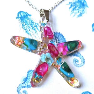 Puede incluir: Un colgante con forma de estrella de mar en una cadena de color plateado, hecho de resina transparente incrustada con piedras pulidas de color rosa, azul y verde, pequeñas flores secas y escamas doradas, creando un aspecto colorido y texturizado.