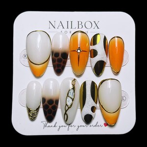 May include: A set of ten oval-shaped press-on nails. The nails feature a variety of designs, including white, orange, and brown gradients, gold accents, and decorative elements. The card reads "NAILBOX FOR U" and "Thank you for your order."