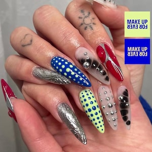 May include: A hand with long, pointed, and elaborately decorated nails. The nails feature various designs, including silver foil, blue with yellow polka dots, red with silver accents, and light green with blue polka dots. Some nails have silver studs and black and white designs. The text "MAKE UP FOR EVER" is visible.