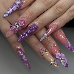 May include: A set of long, pointed acrylic nails in various designs. Some nails feature a light pink base with purple accents, while others have 3D floral and bubble-like embellishments. Rhinestones add sparkle.