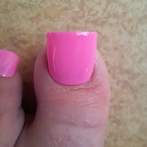 Könnte beinhalten: Nahaufnahme eines Zehs mit leuchtend pinkfarbenem Nagellack. Der Lack hat eine einfarbige, glänzende Farbe und bedeckt den gesamten Nagel. Die Haut um den Zeh ist sichtbar, und der Hintergrund ist eine hellbraune Oberfläche.