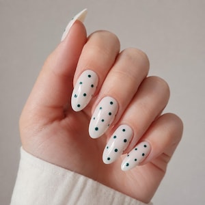 Può includere: Unghie a forma di mandorla con una base lucida color bianco sporco decorata con piccoli pois verde scuro. Le unghie sono curate con una punta arrotondata e liscia, mostrando un motivo pulito e uniforme su tutta la superficie dell'unghia.