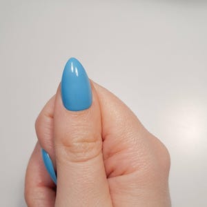 May include: A hand displaying a light blue, almond-shaped artificial nail. The nail has a glossy finish and reflects light. Other nails are also painted light blue. The background is a plain white surface.