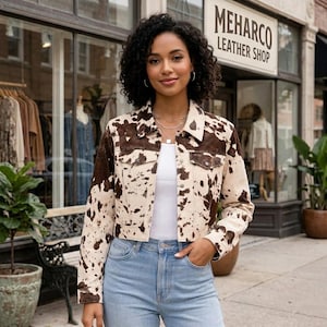 May include: A cropped jacket featuring a brown and cream cow print pattern, a standard collar, long sleeves, and two chest pockets with button closures. The jacket is worn over a plain white top and light blue denim jeans.
