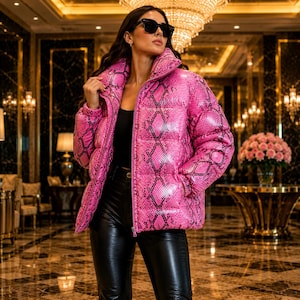May include: A bright pink puffer jacket featuring a black snakeskin print pattern, a high collar, and a front zipper closure. The jacket is worn over a black top and black trousers, paired with dark-tinted sunglasses for a complete look.