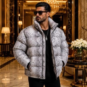 May include: A puffer jacket featuring a grey and white snakeskin pattern with a high collar and a front zipper closure. The jacket is worn over a black shirt and is paired with a silver chain necklace and dark sunglasses with black frames.