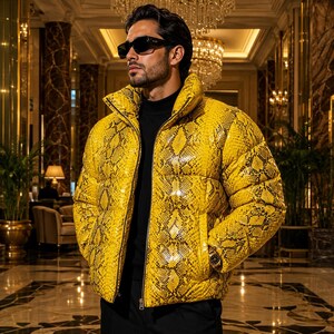 May include: A bright yellow puffer jacket featuring a black snakeskin pattern, a high collar, and a front zip fastening. The jacket is worn over a black mock neck shirt, paired with black sunglasses and a metallic wristwatch on the left wrist.