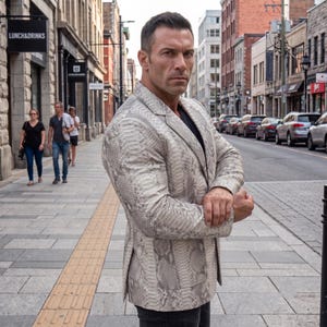 May include: A light grey and white blazer featuring a detailed snakeskin pattern throughout. The garment has a notched lapel, long sleeves, and two front patch pockets. The blazer is worn over a plain black shirt and paired with dark trousers.
